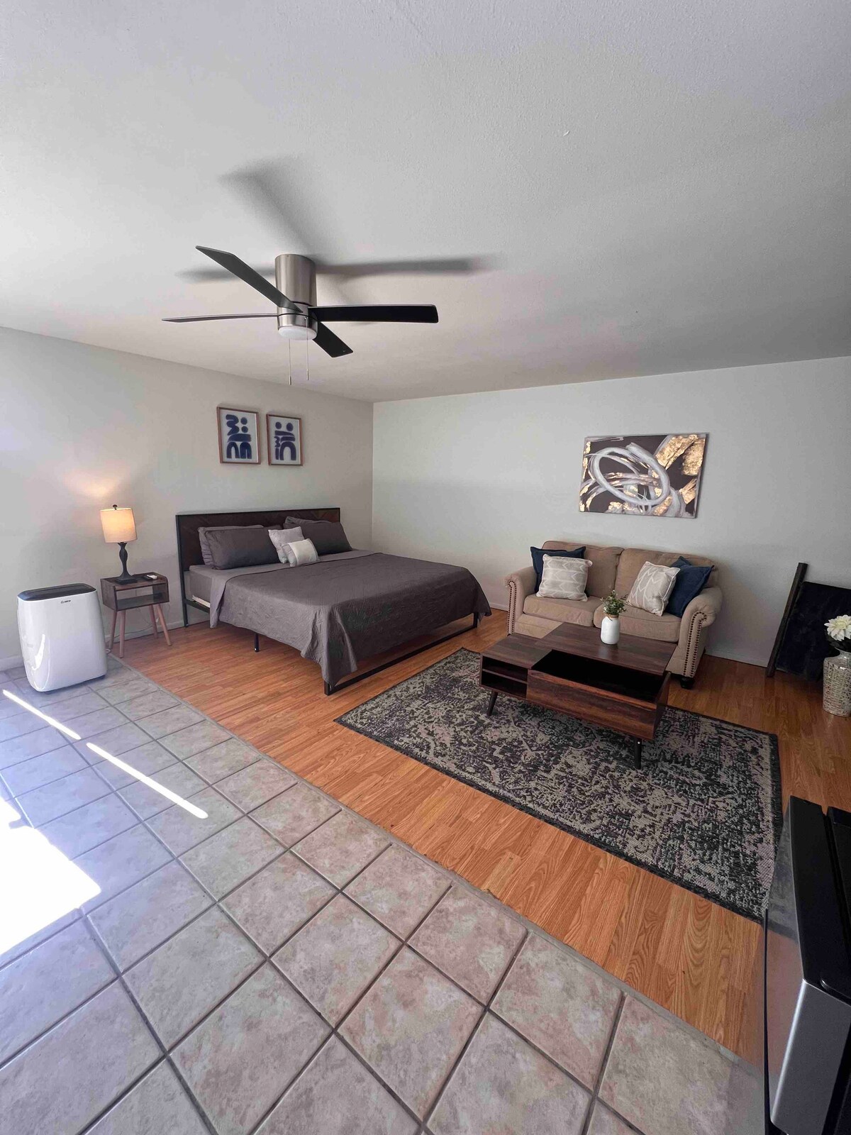 A private suite features a spacious bed topped with a dark gray coverlet, complemented by a small seating area with two armchairs. A coffee table sits in front of the seating, and a minimal design is highlighted by soft lighting. The floor combines tiles and laminate wood.