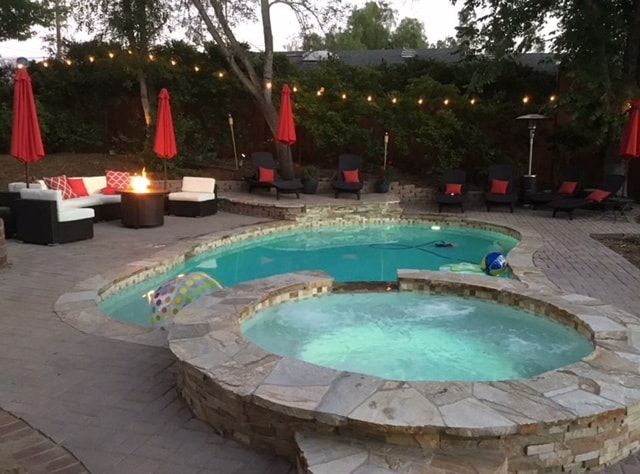 A shared outdoor area features a pool and a jacuzzi, both surrounded by a stone deck. Lounge chairs are positioned nearby under red umbrellas, and a fire pit is visible, inviting relaxation. Soft lighting illuminates the space, creating a welcoming ambiance for guests.