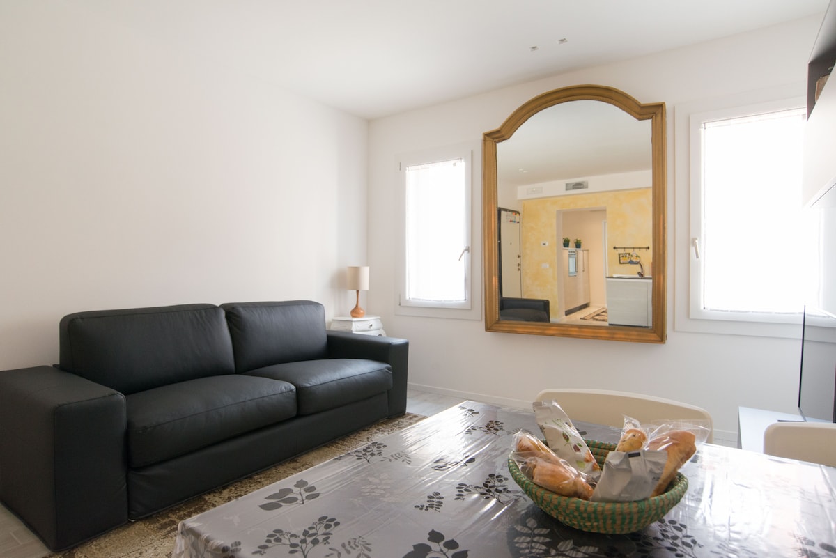 A cozy living area features a modern black sofa positioned next to a light-colored table. A bowl of bread sits at the center of the table. Natural light enters through two windows, with a large decorative mirror on the opposite wall reflecting the space.