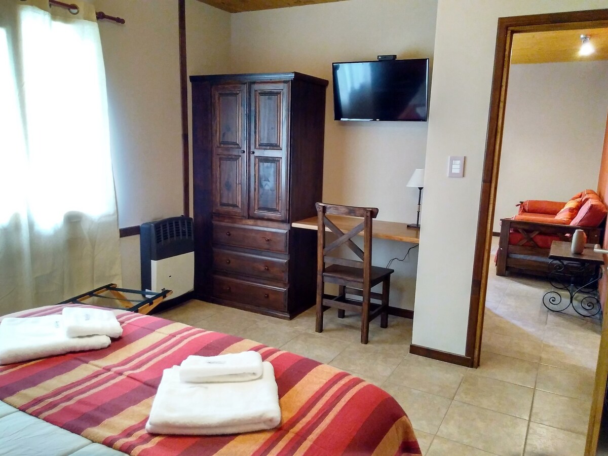An inviting bedroom features a spacious bed covered with neatly arranged white towels. A wooden dresser and a small desk with a chair are positioned against the wall. A flat-screen TV is mounted above the dresser, and natural light filters through the window draped with light curtains.