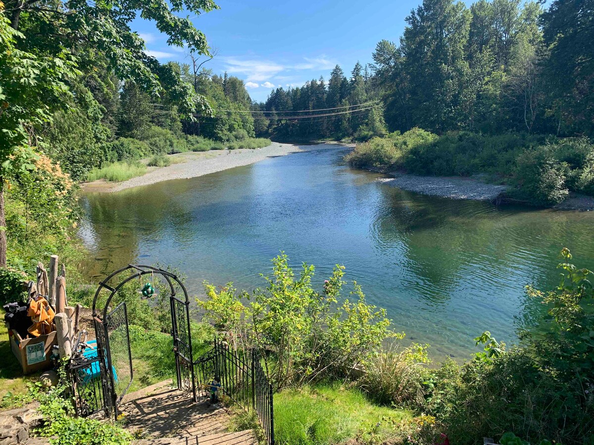 A serene view of the crystal-clear Nanaimo River is presented, framed by lush greenery and trees. The calm water reflects the blue sky above. A winding path descends toward the river, accompanied by an ornamental archway, suggesting a tranquil access point to nature.