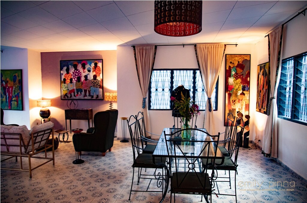 A spacious living area features a glass dining table surrounded by metal chairs. Colorful artwork adorns the walls, with a floor lamp providing soft light. Large windows are dressed with curtains, allowing natural light to brighten the room's eclectic design.
