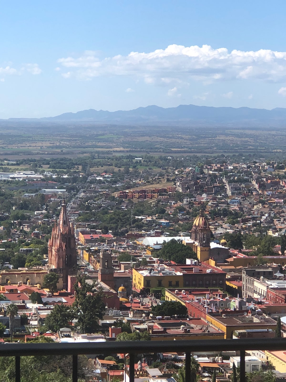A panoramic view of San Miguel de Allende captures the vibrant cityscape, showcasing colorful buildings and intricate architecture. The scene is framed by distant mountains and a clear blue sky, creating a serene backdrop against the bustling town below.