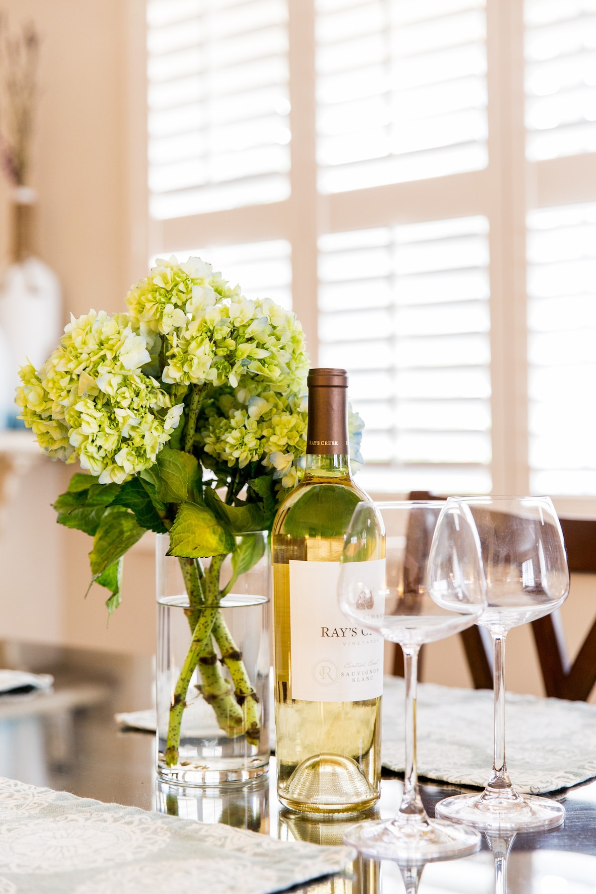 A dining table is set with a clear glass vase holding fresh green hydrangeas. A bottle of white wine stands beside the vase, along with two elegant wine glasses, ready for serving. Light streams through window shutters, enhancing the inviting table setting.