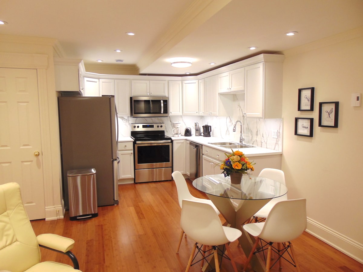 A well-equipped kitchen is presented with modern stainless steel appliances and ample white cabinetry. A round glass dining table with white chairs is positioned in the center, complemented by a colorful floral arrangement. Natural light brightens the space, enhancing the warm wooden flooring.