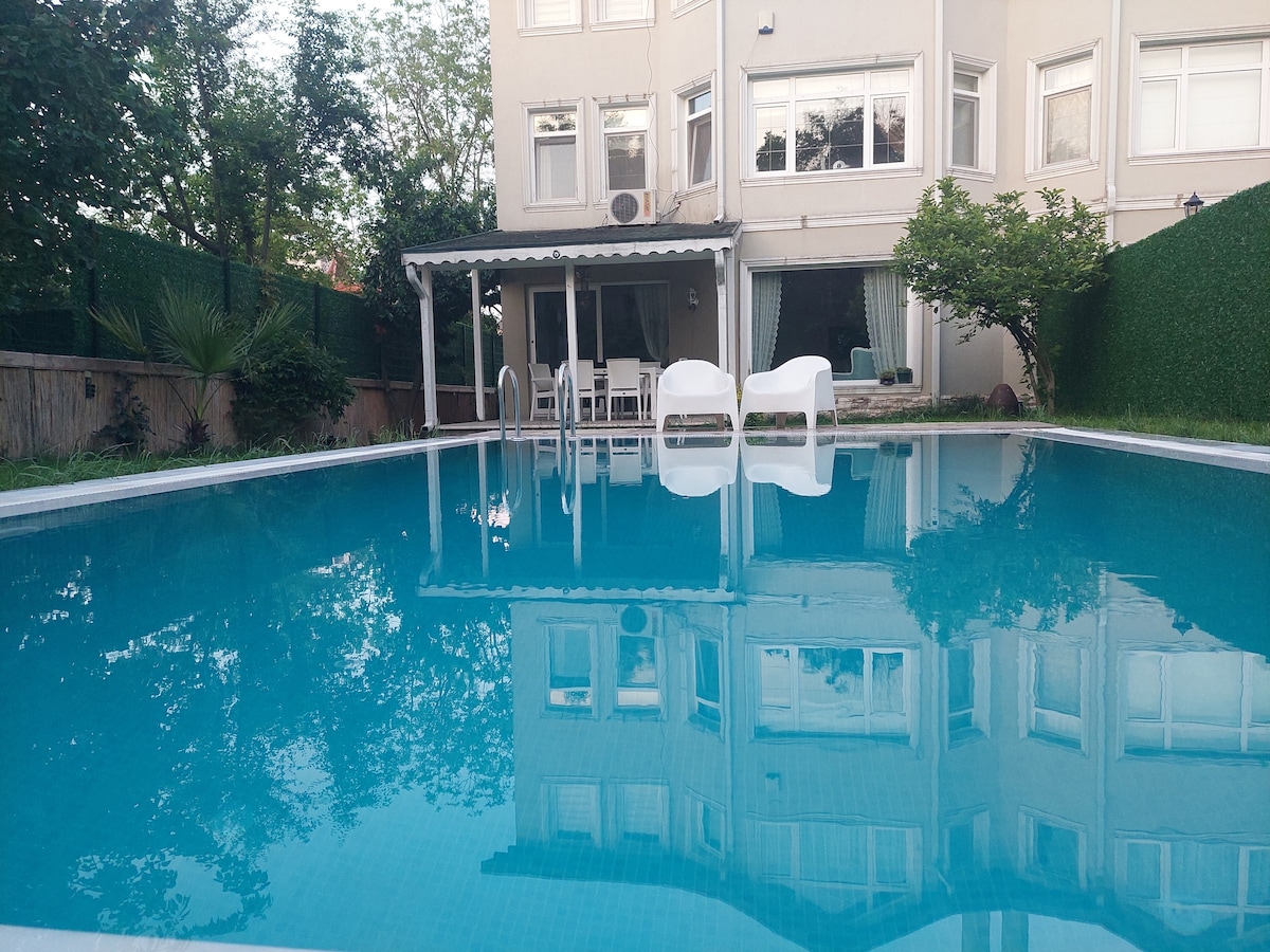 A clean, blue swimming pool reflects the building and surrounding trees. Outdoor furniture, including white chairs and a table, is arranged near the pool. Lush greenery lines the perimeter, creating a peaceful outdoor space.