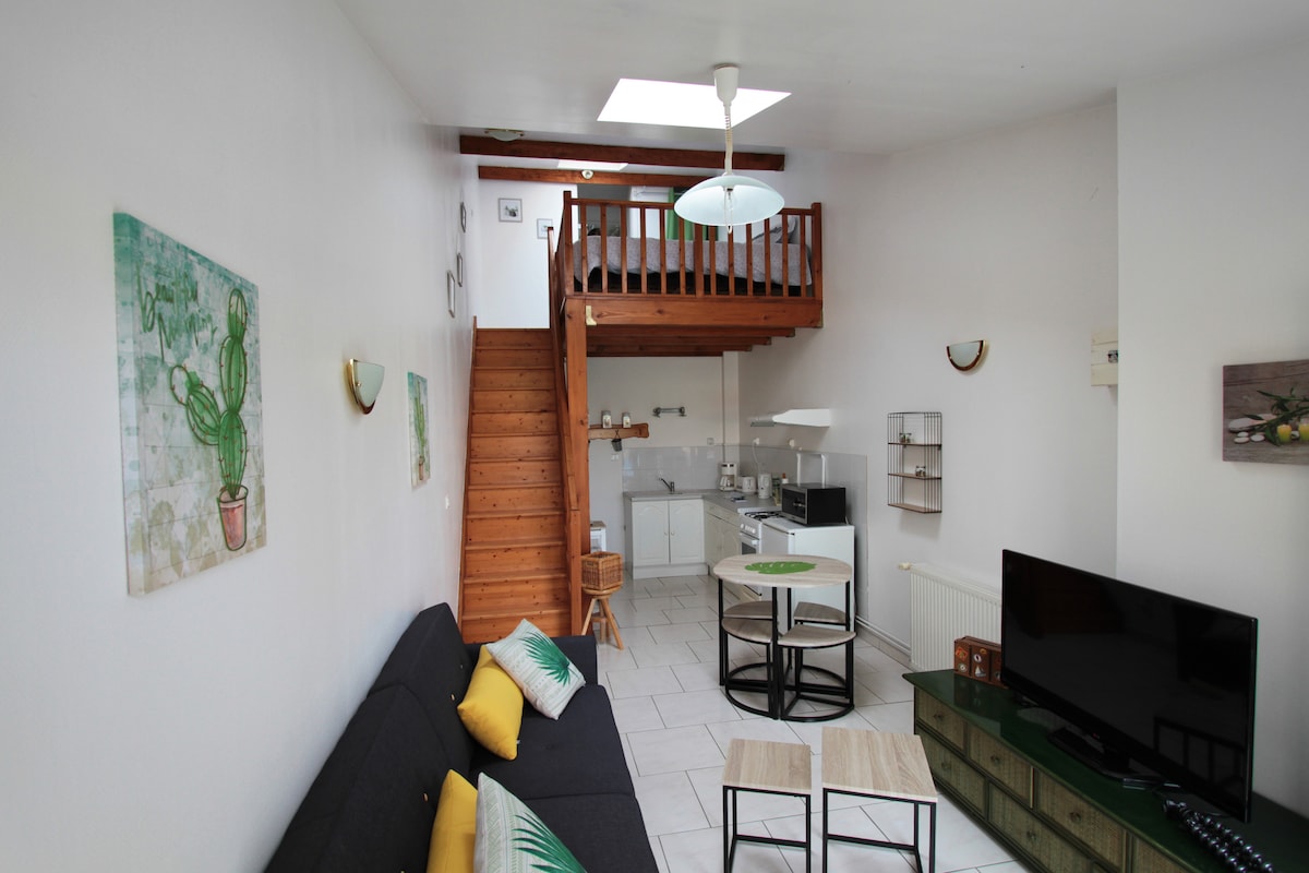 A bright and airy studio features a wooden staircase leading to a lofted area. A cozy seating arrangement includes a dark sofa with decorative pillows. A dining table is set for two, and a television stands on a console beneath the loft. The kitchen area is visible in the background.