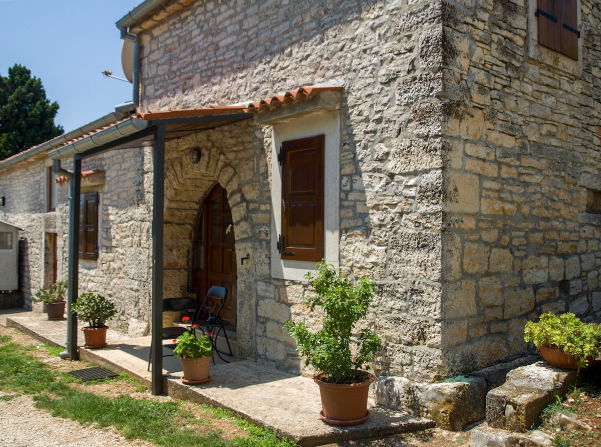 The exterior of a stone house reveals rustic architecture typical of the region. An arched entrance is framed by wooden windows, with potted plants adorning the entrance. A blue chair is positioned by the door, surrounded by a gravel pathway leading to a green lawn.