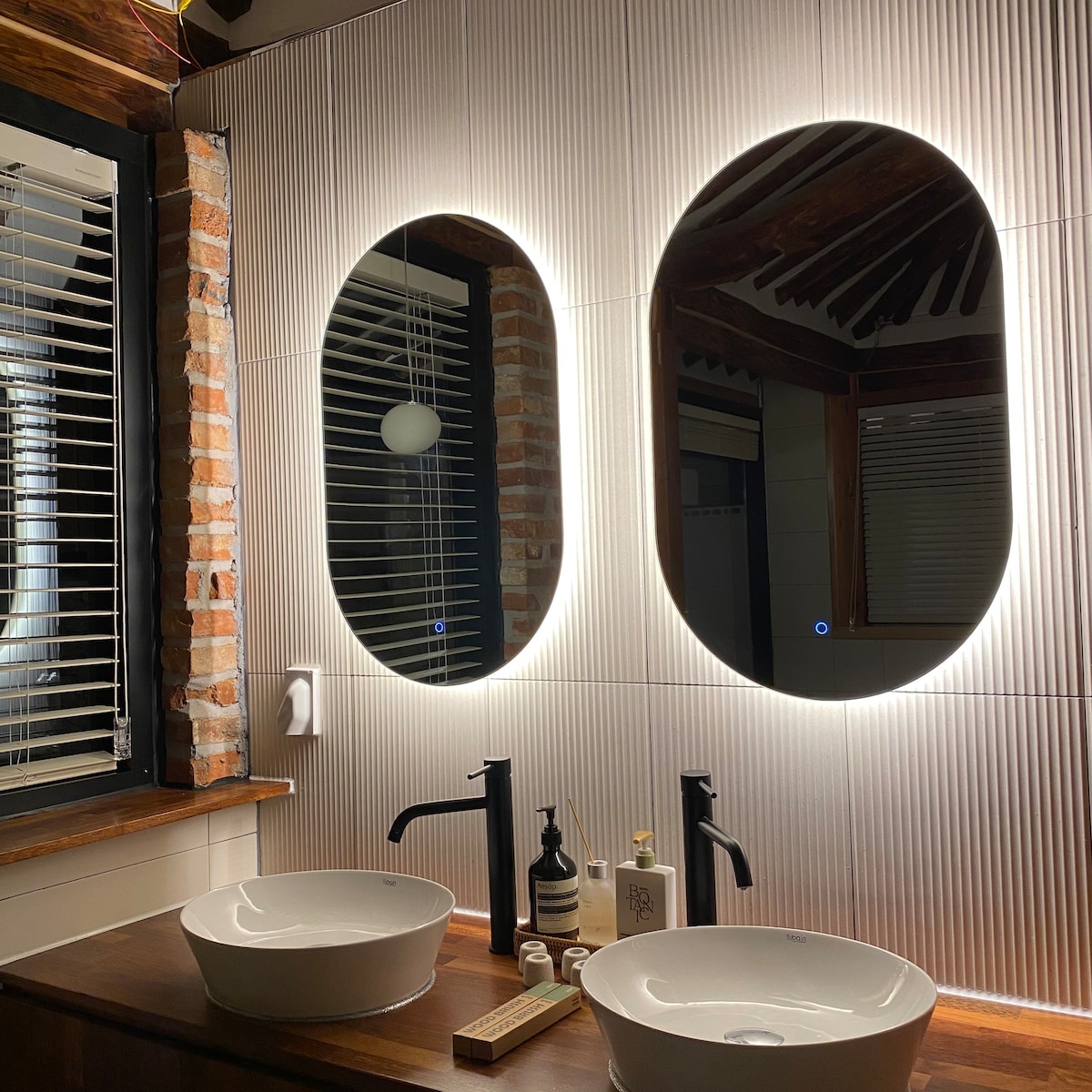 A modern bathroom features two oval mirrors with integrated lighting, reflecting a soothing ambiance. Below, two round sinks are placed on a wooden countertop, accompanied by stylish toiletries and a wooden soap dish. Textured walls and exposed brick details enhance the design.