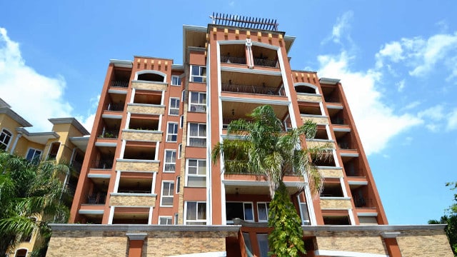 The exterior of a multi-story building is showcased against a clear blue sky. The structure features a combination of stone and stucco elements, with balconies accentuated by decorative railings. Lush greenery surrounds the base, enhancing the inviting atmosphere.
