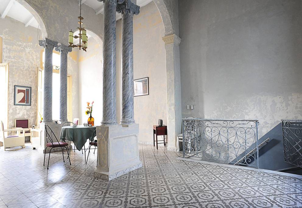 An elegant colonial-style living area features high ceilings supported by tall columns. A dining table is set with chairs, and natural light filters through large windows. Decorative floor tiles create a patterned visual that complements the neutral walls and simple furnishings.