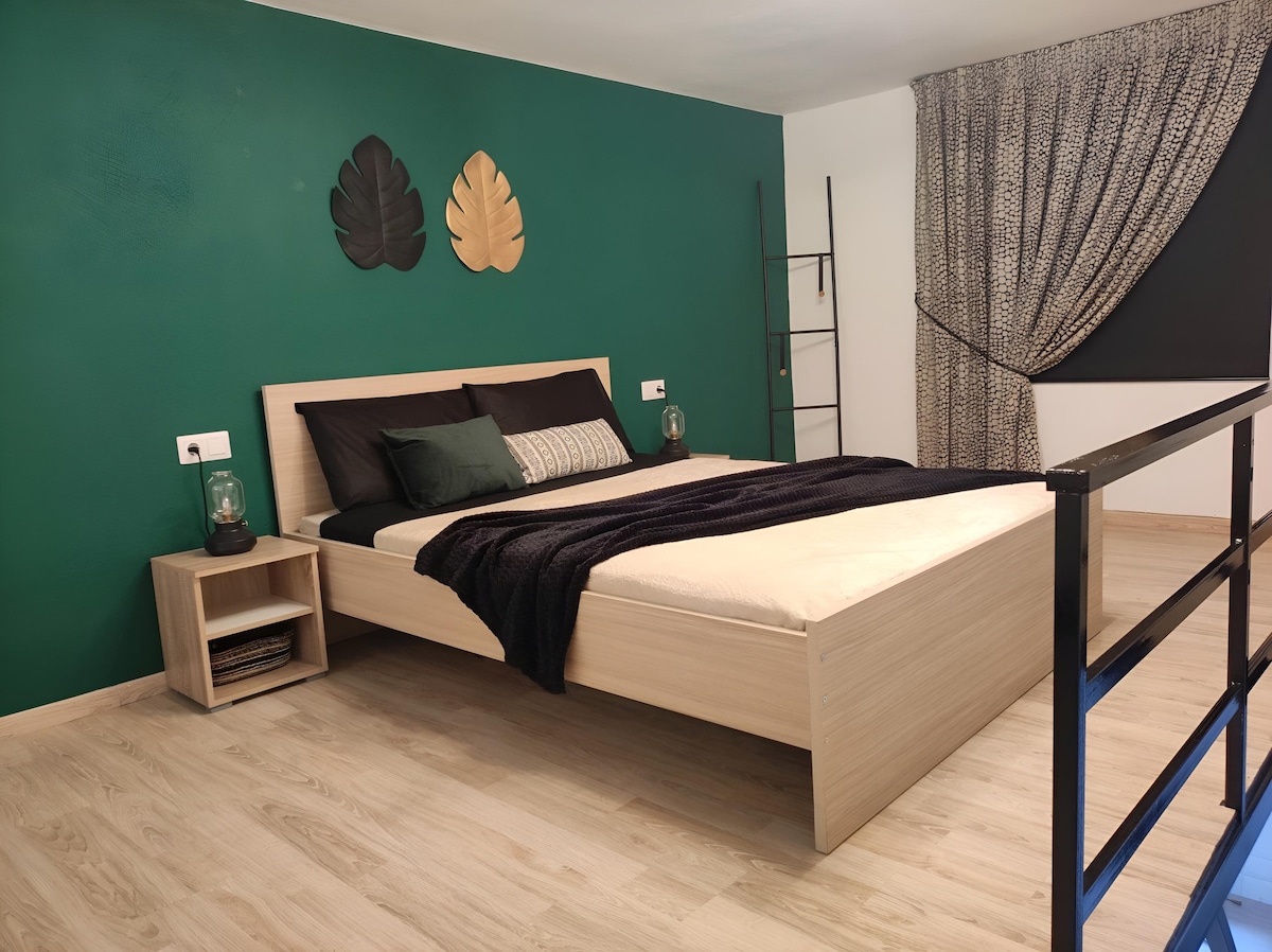 A cozy sleeping area features a spacious bed with neutral bedding against a vibrant green wall. Two decorative leaf wall pieces enhance the room's style. A tall ladder shelf stands nearby, while a curtain partially covers a window, allowing soft light into the space.