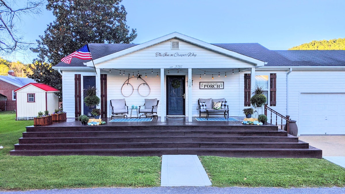 The front of The Inn on Creeper Way is showcased with a welcoming porch featuring two seating arrangements, adorned with blue cushions, surrounded by landscaped greenery. An American flag is displayed, while string lights enhance the inviting appearance of the entrance.