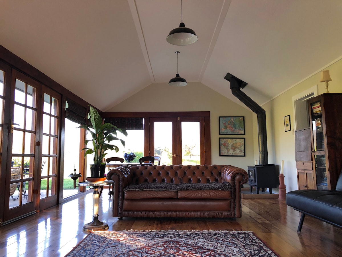 A spacious living area features a classic brown leather chesterfield sofa, complemented by large windows allowing natural light. A cozy wood-burning stove is visible, along with a decorative area rug. Indoor plants are placed to enhance the inviting ambiance of the room.