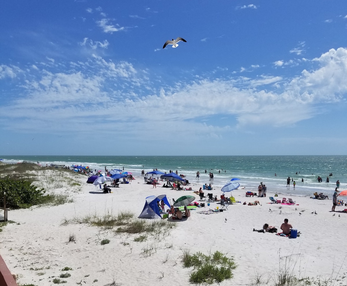 A sandy beach is populated with umbrellas and beachgoers enjoying the sun. Gentle waves can be seen lapping at the shore, while a blue sky dotted with clouds stretches overhead. The scene showcases a peaceful coastal atmosphere.
