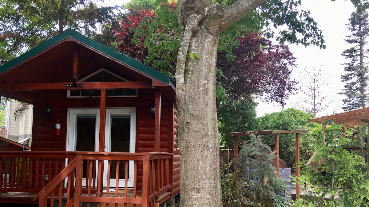 A charming cabin is framed by lush greenery and colorful trees, showcasing a wooden deck that offers outdoor seating. Large windows provide views into the inviting interior, complemented by a well-maintained outdoor space featuring additional landscaping.