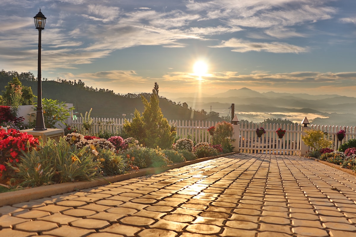 A well-maintained pathway made of smooth cobblestones leads through vibrant flower beds towards a white picket fence. The sun rises over distant mountains, casting a soft glow on the landscape and creating an inviting atmosphere.