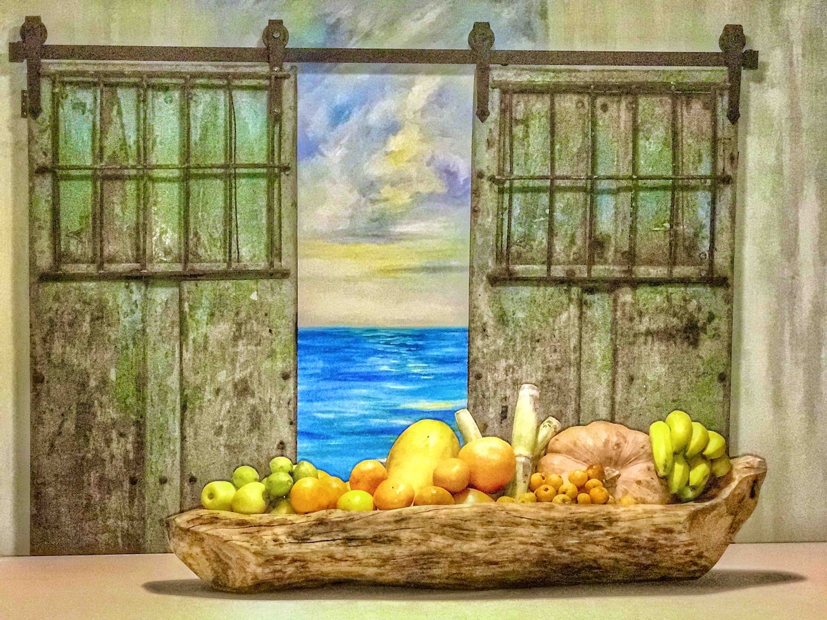 A decorative wooden bowl filled with a variety of colorful fruits sits prominently in front of a mural depicting a serene seascape. Behind the bowl, sliding barn doors open to reveal an ocean view, enhancing the room's lively ambiance.