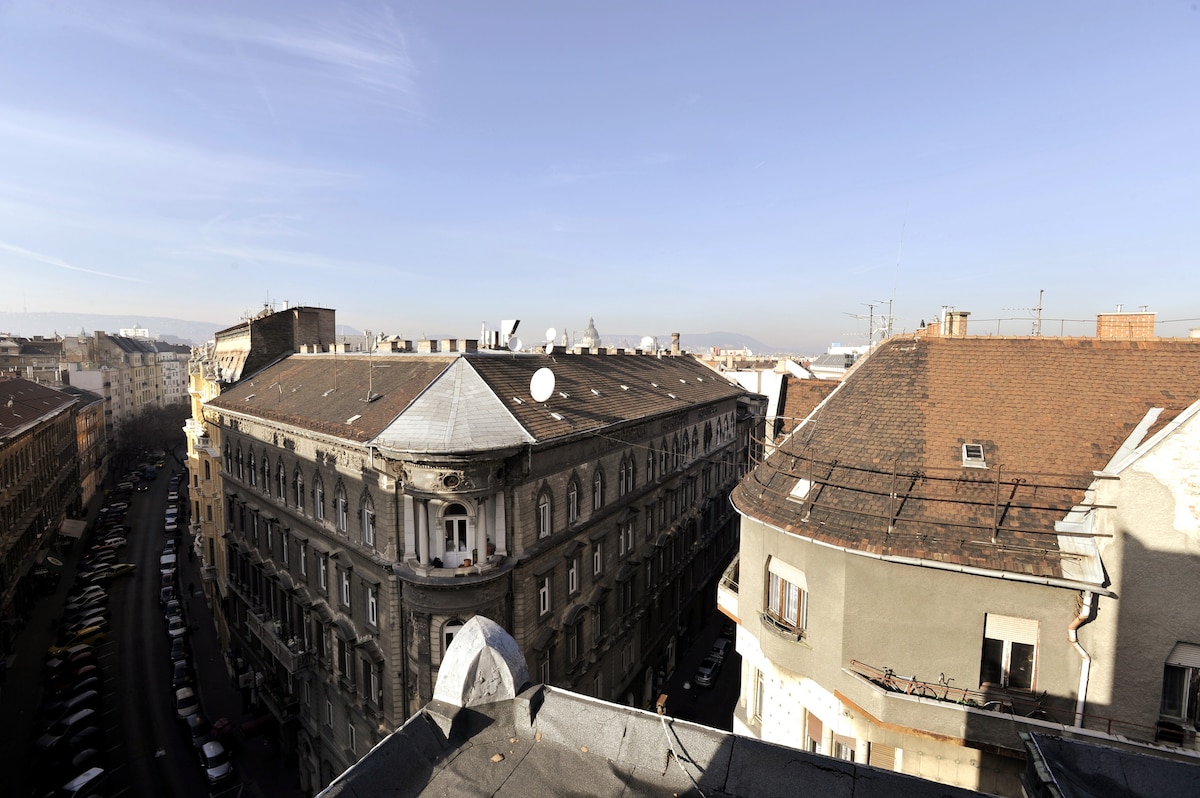 A panoramic view shows the rooftops of Budapest’s architecture, with multiple buildings featuring varying designs. Satellite dishes dot the rooftops, while a clear blue sky stretches overhead, highlighting the urban scene below.