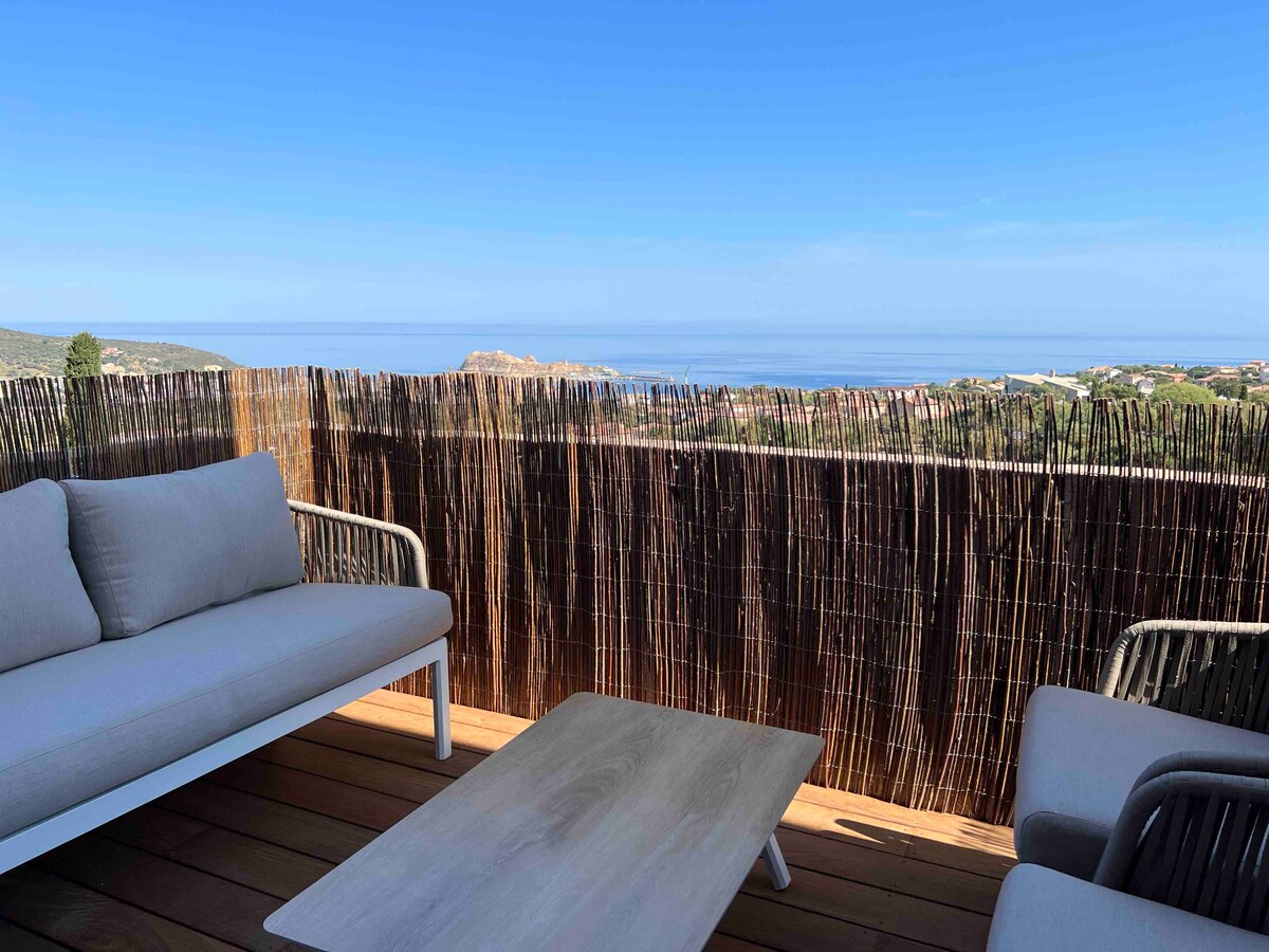 A private terrace is featured, furnished with a light-colored sofa set and a wooden coffee table. A natural privacy screen provides separation while offering views of the sea and surrounding landscape. Clear blue skies encompass the area, enhancing the outdoor setting.