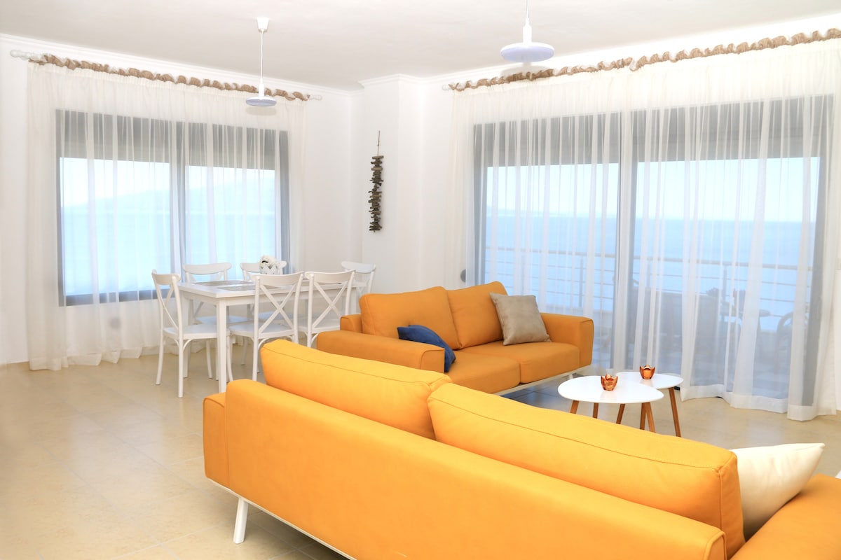A bright living area features large windows offering a sea view, complemented by sheer curtains. A modern orange sofa is arranged alongside a small round table. A dining table with white chairs is positioned nearby, providing an inviting space for meals and gatherings.