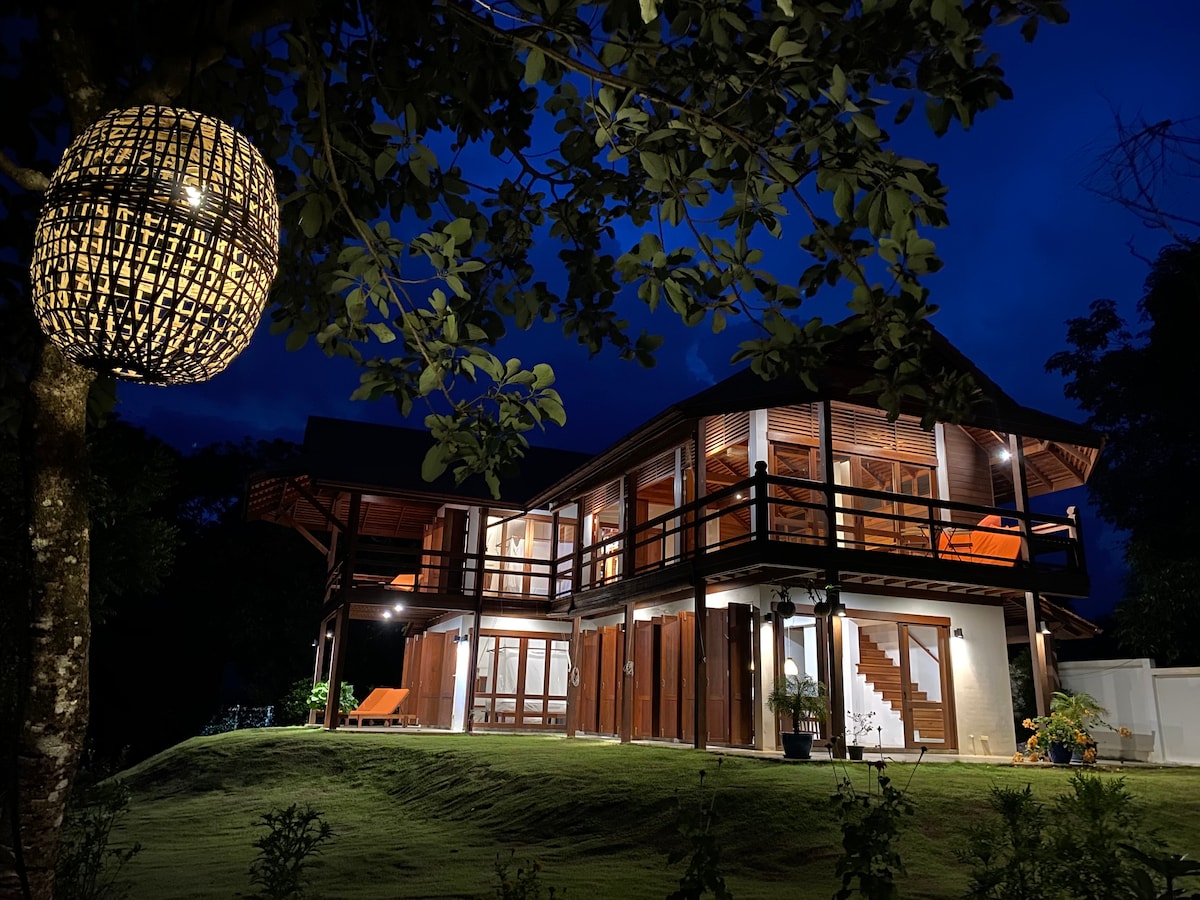 The villa is illuminated at night, showcasing its modern architectural features. Large windows and balconies are visible, surrounded by lush greenery. A decorative lantern hangs from a tree branch, adding to the serene atmosphere of the outdoor environment.