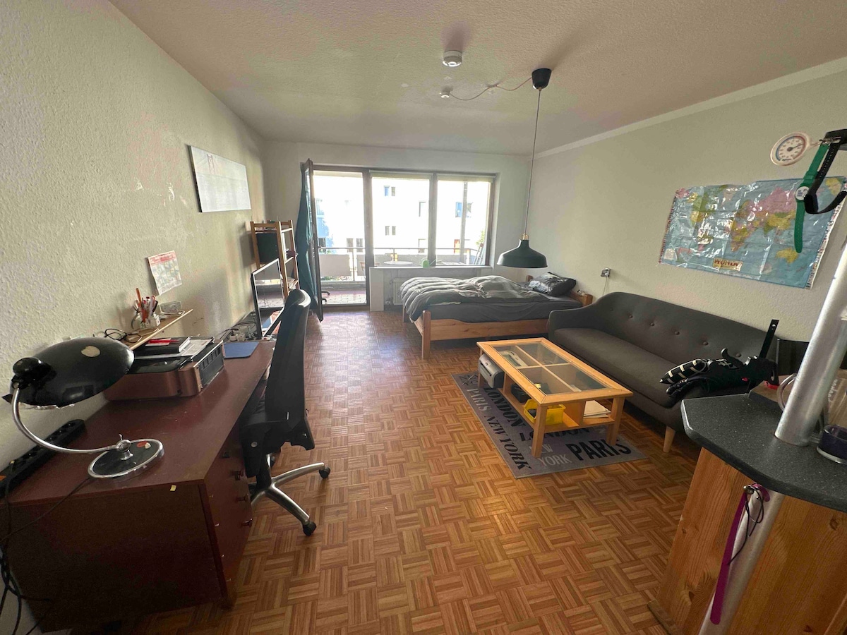 The living space features a functional layout with a cozy sofa, a coffee table, and a workstation. A spacious bed lies beside a large window, providing natural light. A balcony is visible through the doors, and the floor displays a patterned layout of wooden tiles.