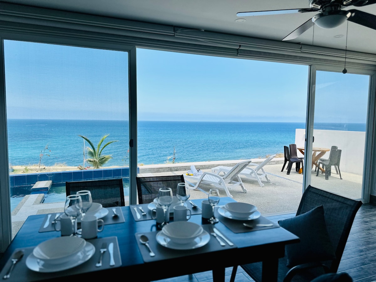 A dining table is set with plates, glasses, and cutlery, positioned near large sliding glass doors. The ocean is visible in the background, with a patio featuring lounge chairs and a pool just outside. Bright sunlight illuminates the space.