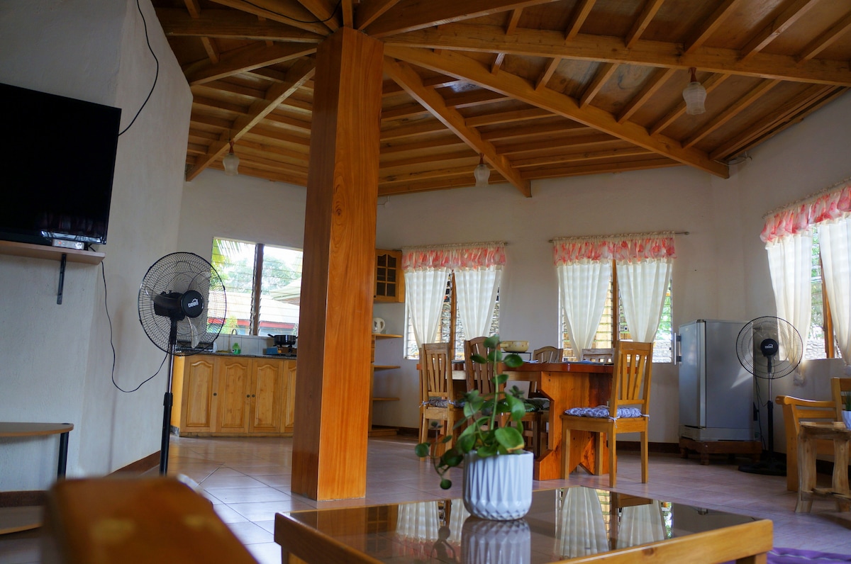 An open living area is highlighted by wooden beams and natural light filtering through sheer curtains. A dining table surrounded by wooden chairs complements a kitchenette, with a refrigerator visible nearby. Decorative plants add a touch of greenery to the space.