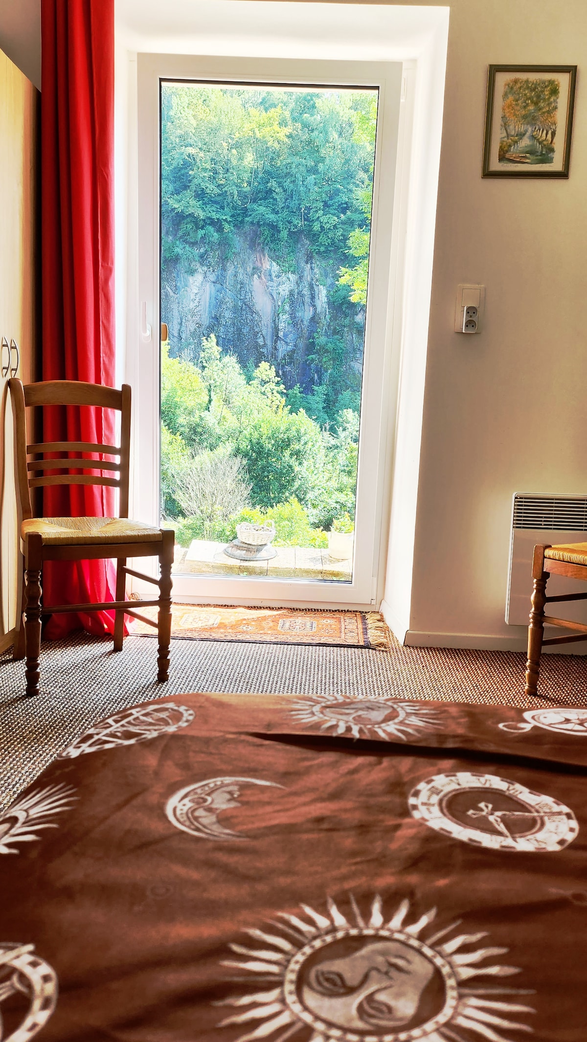 Natural light filters into the room through the large glass door, providing a view of the lush greenery and a cliff outside. A brown patterned blanket covers the bed, while chairs are positioned nearby, maintaining a simple and inviting atmosphere.