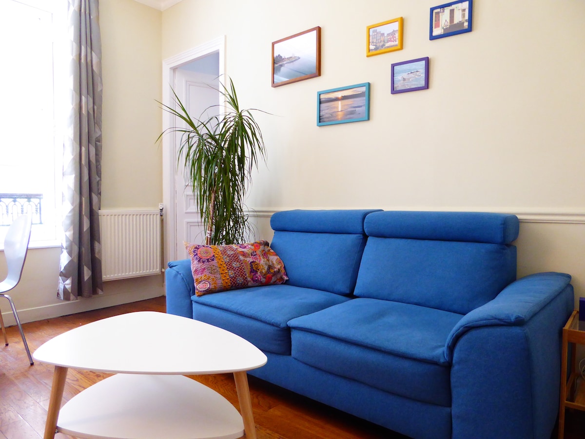 A comfortable blue sofa is positioned in the living area, accompanied by a decorative pillow. A small white coffee table sits in front. A tall green plant adds a touch of nature, while framed photographs adorn the light-colored wall, enhancing the space's visual interest.
