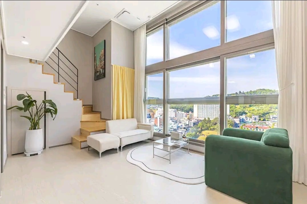 A bright living area features large windows offering expansive city and mountain views. A green sofa is positioned beside a sleek glass coffee table, with a white couch across. A wooden staircase leads to an upper level, complemented by soft curtains framing natural light.
