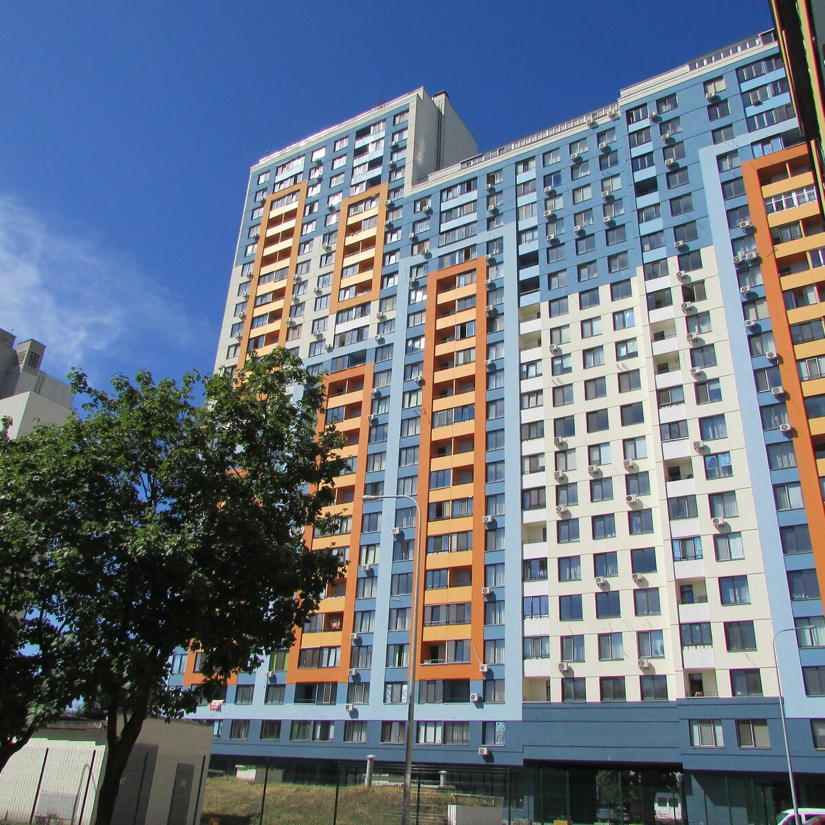 A modern high-rise building is depicted, featuring a mix of blue and orange façade elements. Large windows provide ample natural light, while a green tree is visible in the foreground, adding a touch of nature to the urban setting.