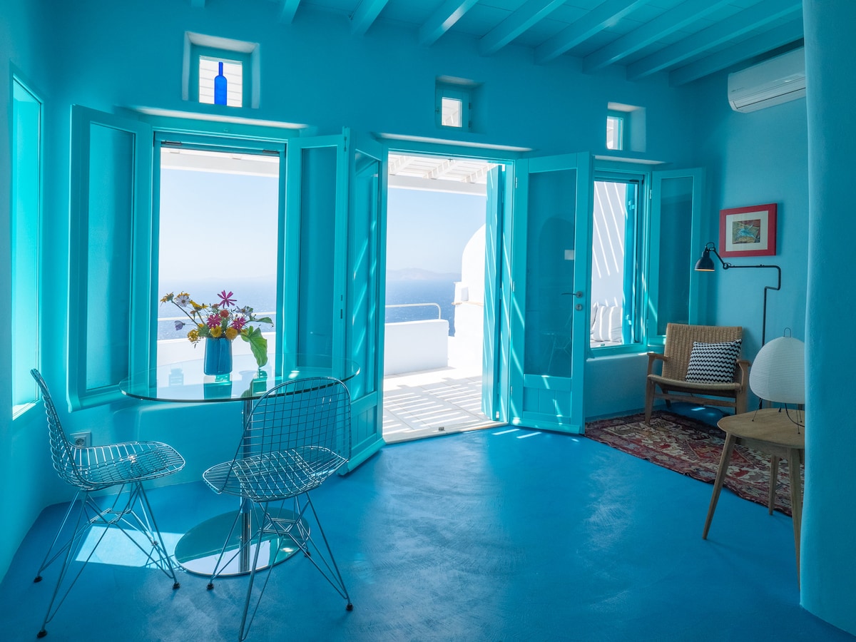 A bright living area features vibrant blue walls and flooring, illuminated by natural light entering through large open windows. A small dining table is set against the window, accompanied by two wire chairs. A cozy reading corner includes a woven chair and a modern lamp.