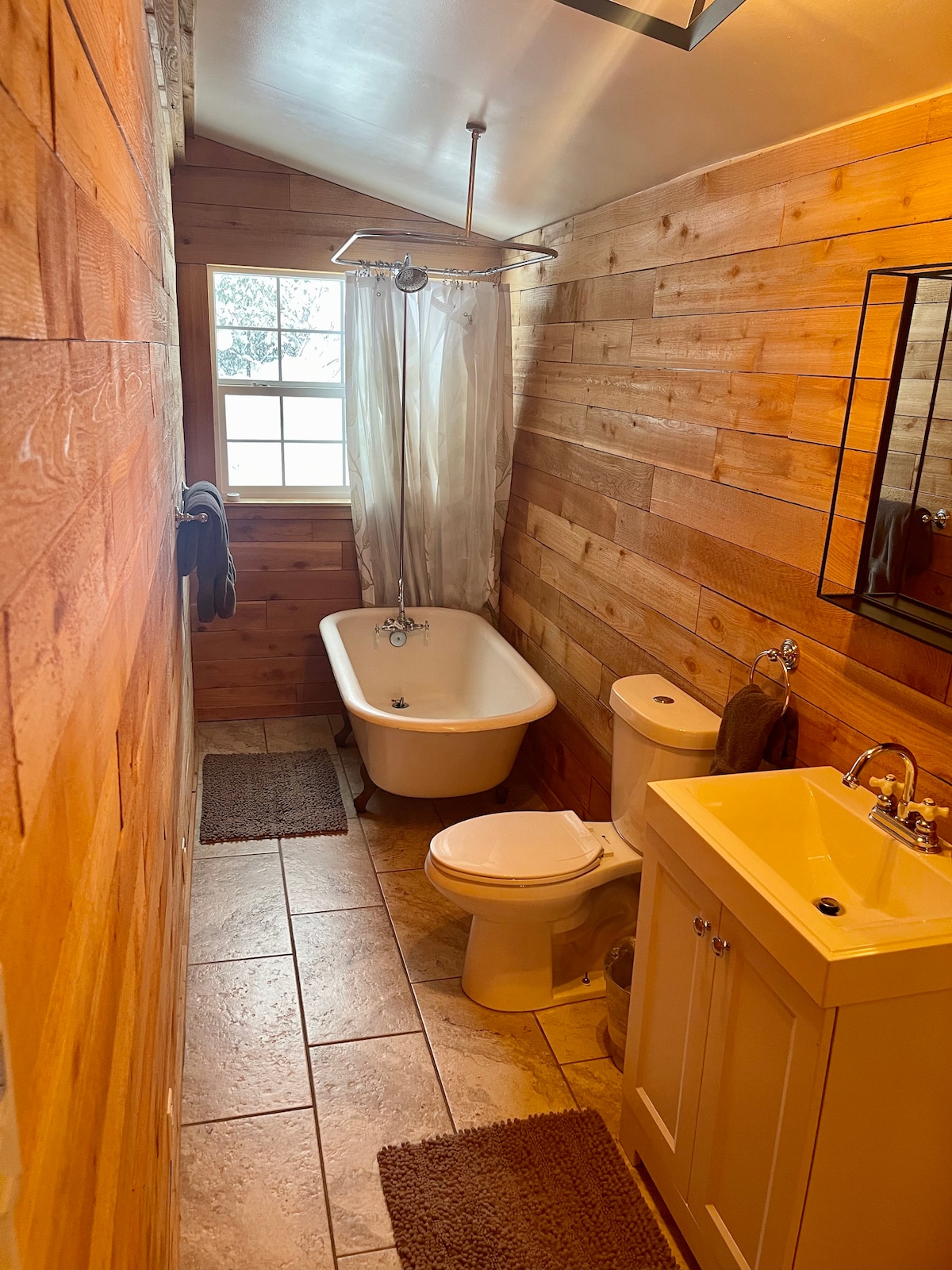 A bathroom features rustic wooden walls and a combination of tile and stone flooring. A freestanding bathtub is set beside a large window, while modern fixtures, including a sink and toilet, are present. Soft rugs provide comfort underfoot.