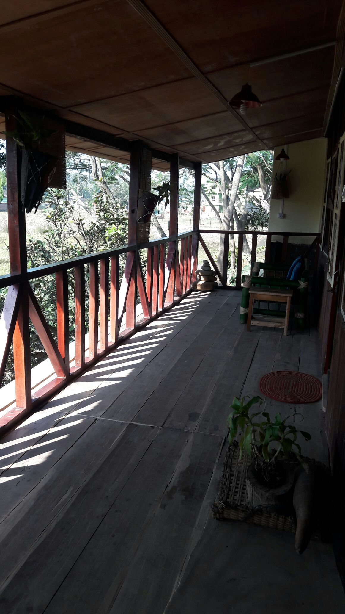A spacious porch features wooden railings and a polished floor, adorned with potted plants and a woven mat. Natural light casts shadows across the area, enhancing the serene view of the surrounding greenery.