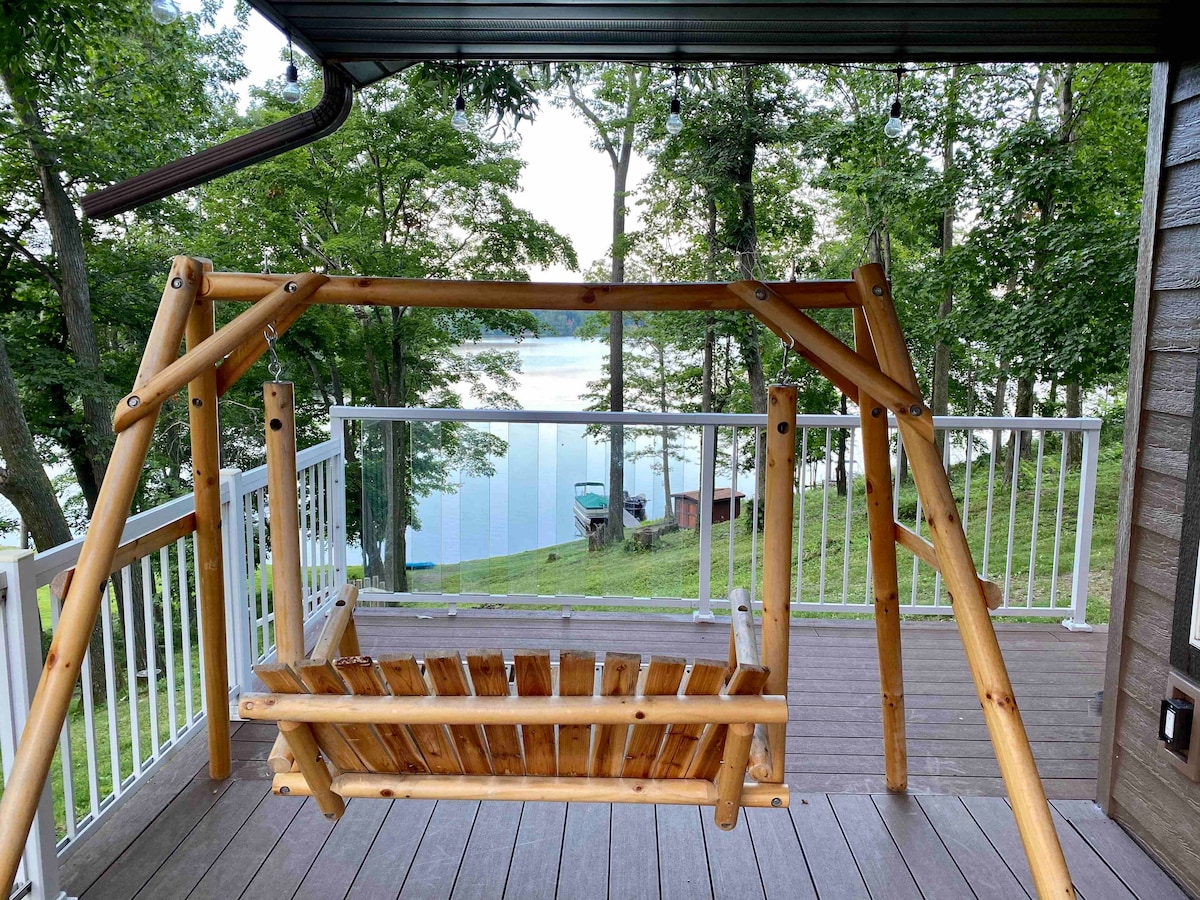 A wooden swing hangs from a sturdy frame on a deck, providing a view of the tranquil lake surrounded by trees. The deck features a railing that offers safety and an unobstructed sightline to the water, enhancing outdoor relaxation.