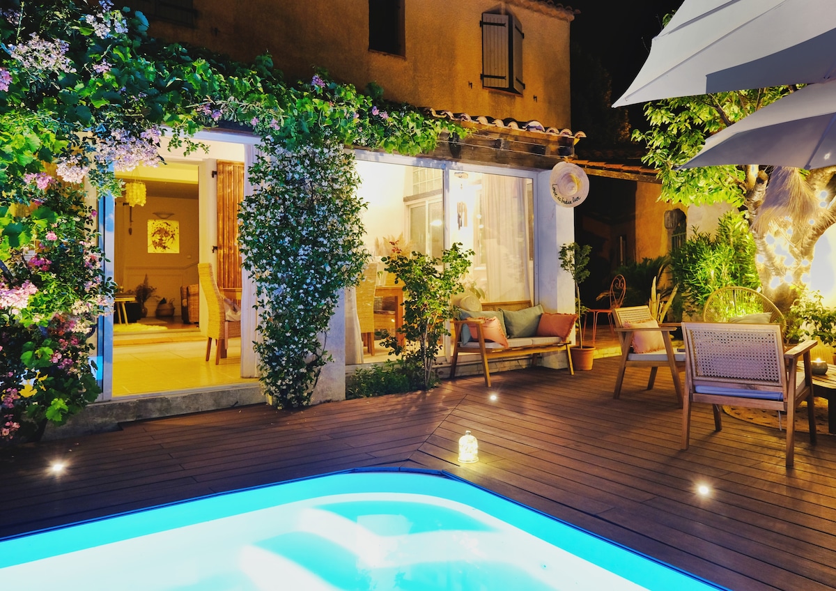 The illuminated swimming pool reflects soft blue lights, enhancing the serene outdoor deck adorned with wooden furniture. Vines and flowering plants gracefully frame the entrance, leading into a warm and inviting living space visible through large windows.