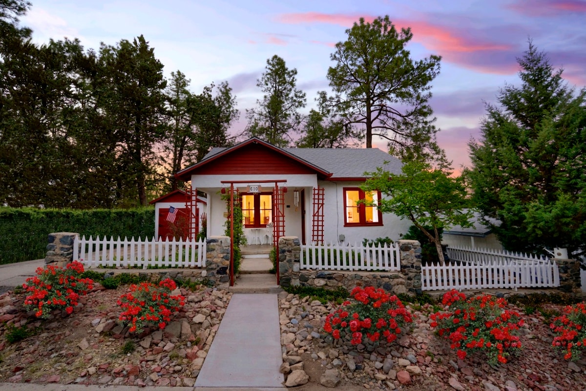 A charming cottage is nestled among tall pine trees, featuring a white picket fence and a flower bed filled with blooming red flowers. The entrance is accentuated by a stone pathway leading to a welcoming porch with large windows that reflect the evening light.