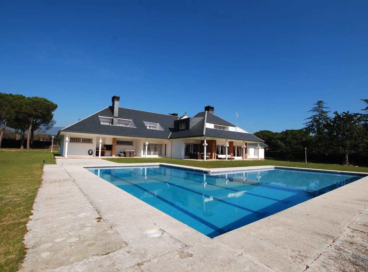 The exterior of a spacious white house is visible, featuring a large pool surrounded by a stone patio. Green lawns stretch around the property, and tall trees provide shade in the background. A clear blue sky complements the tranquil setting.