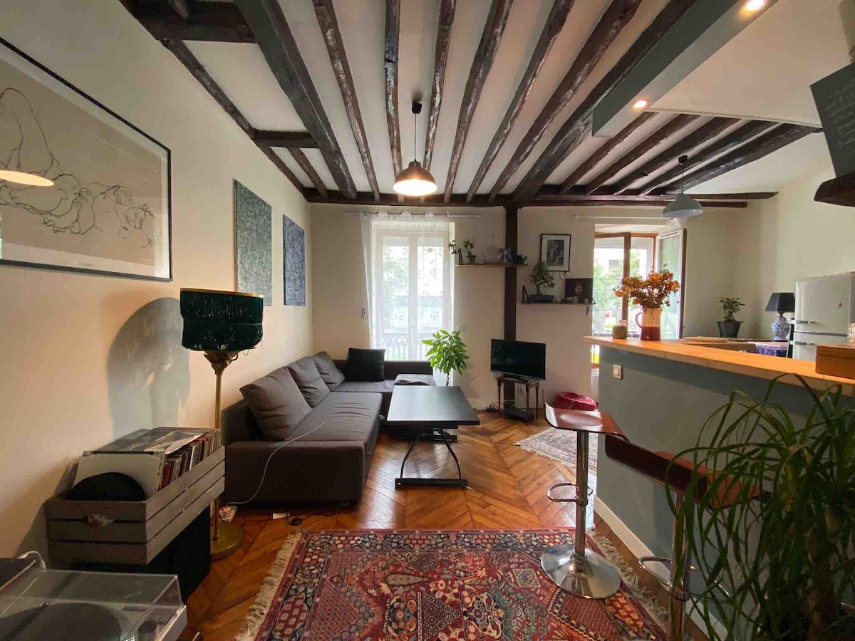 The living area features a spacious sectional sofa facing a modern coffee table, with natural light filtering through a window. Dark wooden beams adorn the ceiling, while framed artwork decorates the walls. A seating area is visible near a bar counter overlooking the kitchen.