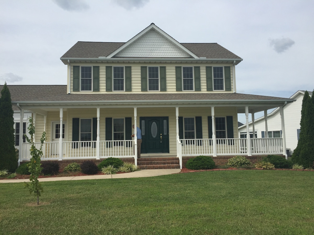 The exterior of a two-story family home is presented, featuring a light yellow facade with white trim. A covered porch stretches across the front, supported by evenly spaced columns. Green lawn surrounds the house, enhancing the tranquil setting, while fluffy clouds drift across the sky.