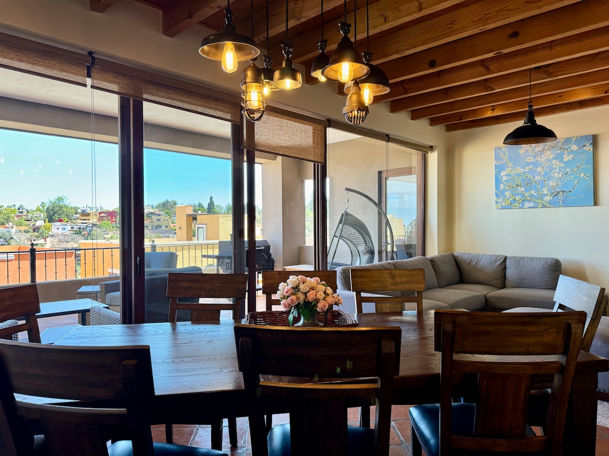 A spacious dining area features a large wooden table surrounded by eight chairs, with a vase of flowers at the center. Large glass doors offer views of the terrace and surrounding landscape, allowing natural light to fill the room. A comfortable seating area is visible in the background.
