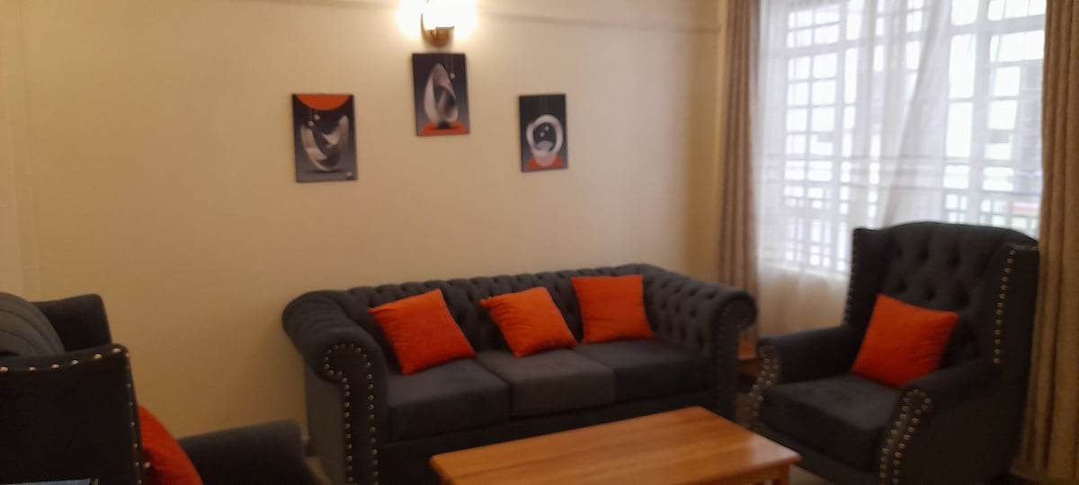 A comfortable living area is furnished with a dark sofa and two armchairs, accented by vibrant orange cushions. A wooden coffee table is situated in the center, while artwork adorns the walls, and natural light filters through sheer curtains.