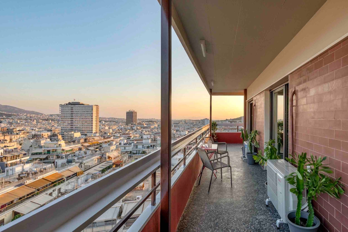 A spacious balcony is showcased, featuring two chairs and small tables positioned against a backdrop of the city skyline. Green plants add a touch of nature. The scene is illuminated by the warm glow of sunset, enhancing the view of the surrounding buildings.
