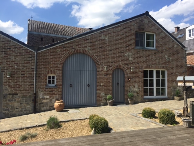 The exterior of the house features a charming façade of red brick and light grey accents. A large arched door and a set of double doors are visible, alongside multiple windows. A landscaped pathway with small shrubs leads up to the entrance, creating a welcoming approach.