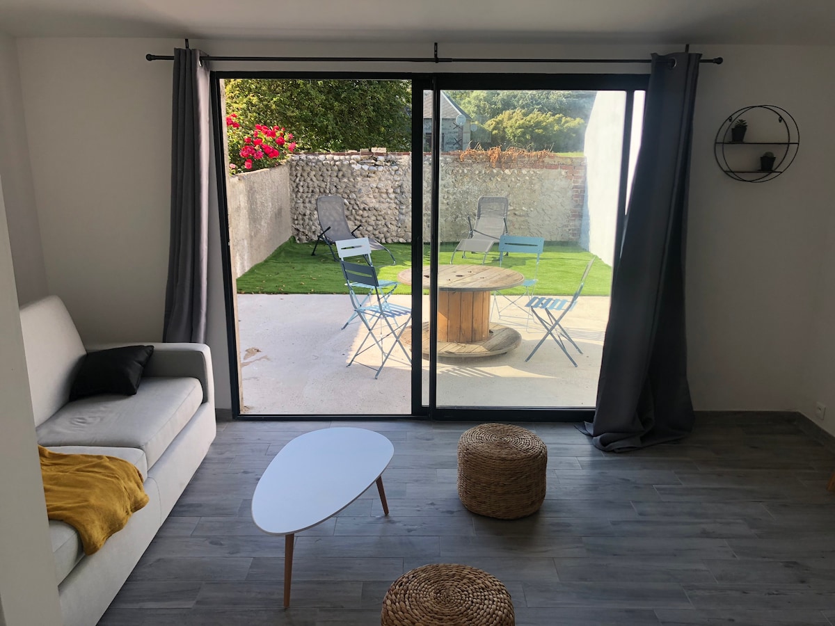 A spacious living area is showcased, featuring a light-colored sofa with a throw, a round coffee table, and natural fiber poufs. Large sliding doors provide a view of the enclosed outdoor space, which includes a circular table with chairs and greenery, inviting natural light into the room.