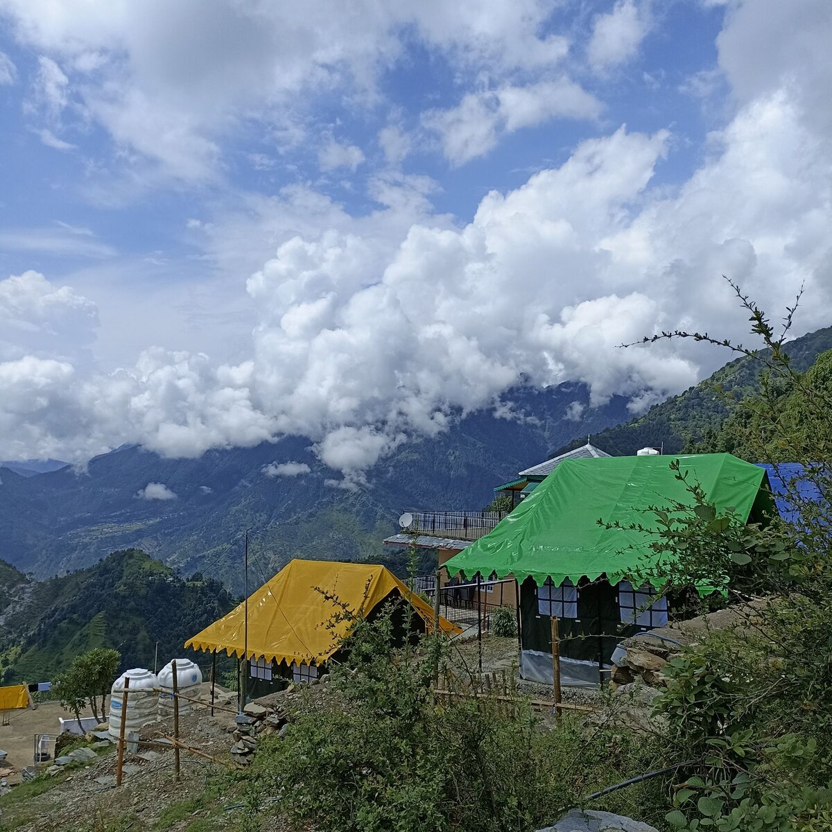 Colorful Swiss tents are set against a backdrop of lush mountains and dramatic clouds. Bright yellow and green tents contrast with the natural landscape, while greenery is visible in the foreground, enhancing the serene outdoor atmosphere.