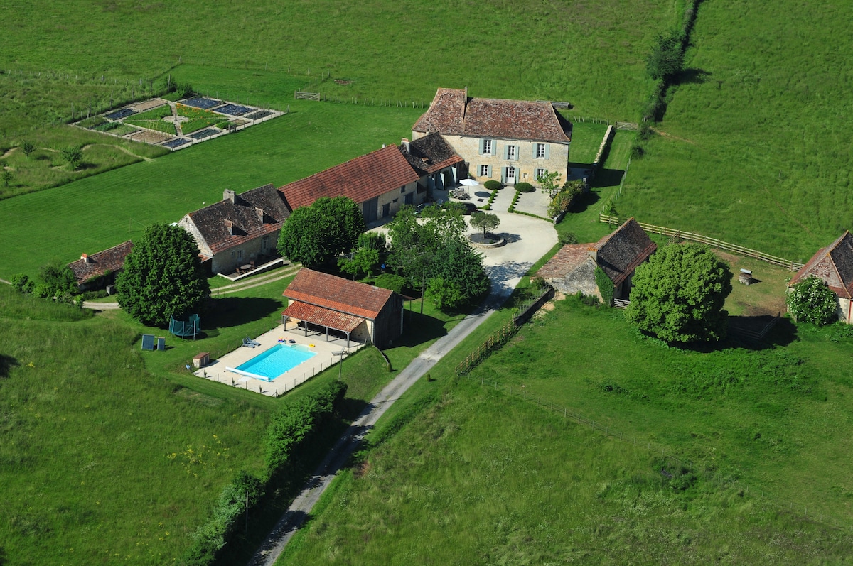 The aerial view captures the expansive property, featuring the restored manor house and a surrounding landscape of lush green fields. A swimming pool is visible, along with shaded outdoor seating areas and well-maintained gardens, illustrating the openness and tranquility of the estate.