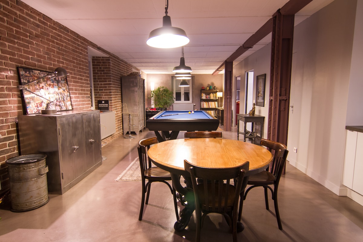 An open living area features a professional pool table at its center, surrounded by warm, industrial-style decor. A round wooden dining table with dark wooden chairs is positioned nearby. Exposed brick walls add character, while modern lighting fixtures provide ample illumination.